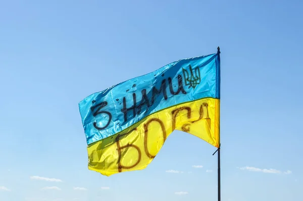Waving Ukrainian yellow-blue flag against a blue sky. On the flag is the coat of arms of Ukraine and the inscription: \