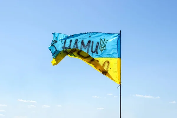 Waving Ukrainian yellow-blue flag against a blue sky. On the flag is the coat of arms of Ukraine and the inscription: \