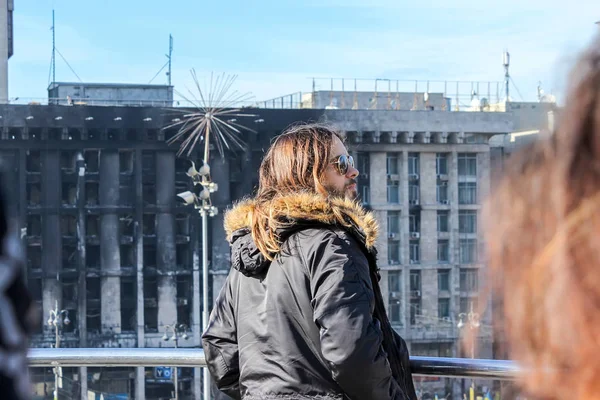 Jared Leto, metal bir çit tarafından ayakta, bir yanmış bina ve Maidan Nezalezhnosti üzerinde Euromaidan zamanlarında bir çadır şehir aşağı bakıyor