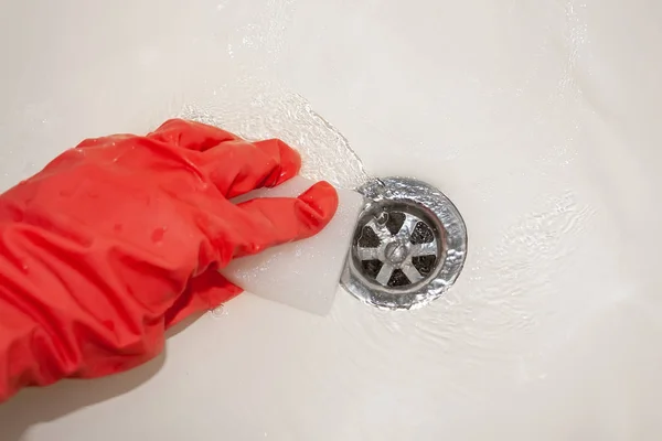 Рука в красной резиновой перчатке с меламиновой губкой массирует белую ванную у сливного отверстия, где течет вода. Выборочный фокус. Вид крупным планом — стоковое фото