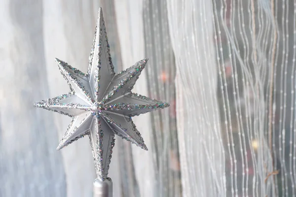 Silver Christmas star with many sparkles against a light textile curtain. Home decoration for a family holiday with copy space. Christmas tree top. Selective focus