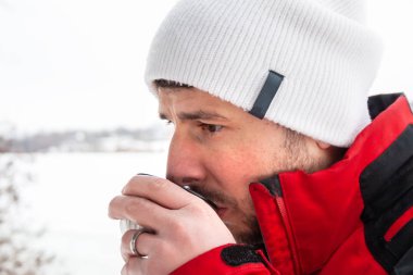 Kırmızı ve siyah ceketli ve beyaz şapkalı yakışıklı sakallı adam donmuş bir nehre karşı metal bir bardaktan sıcak çay içiyor.