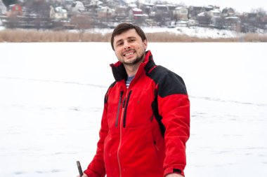 Kırmızı ceketli bir adam gülümsüyor, donmuş bir nehirde duruyor. Nehrin çok ötesinde küçük evler görülebilir