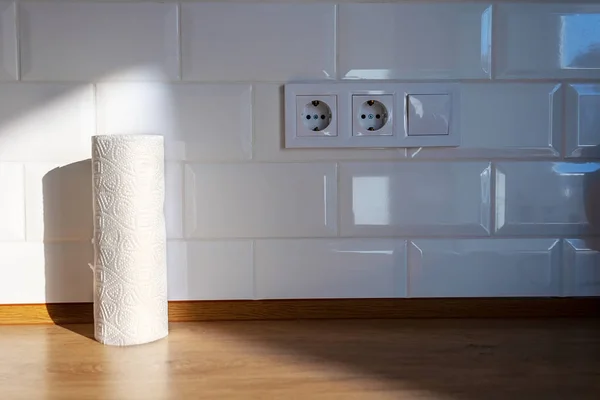 Eine Rolle weiß gemusterter Küchenpapierhandtücher steht in der Sonne auf einer hölzernen Arbeitsplatte vor einer Wand aus weißen Fliesen, daneben stehen zwei europäische Steckdosen und ein Schalter — Stockfoto
