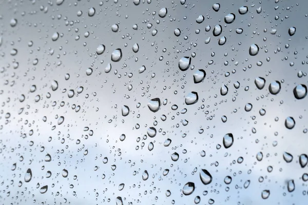 Капли воды на стеклянное окно на размытом голубом и сером фоне неба в дождливый осенний день. Селективный фокус — стоковое фото