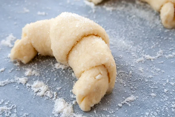 A closeup view of a croissant prepared for baking from a curd dough sprinkled with sugar lying on a parchment baking paper covered with flour. Sweet bagel before loading into the oven — Stock Photo, Image