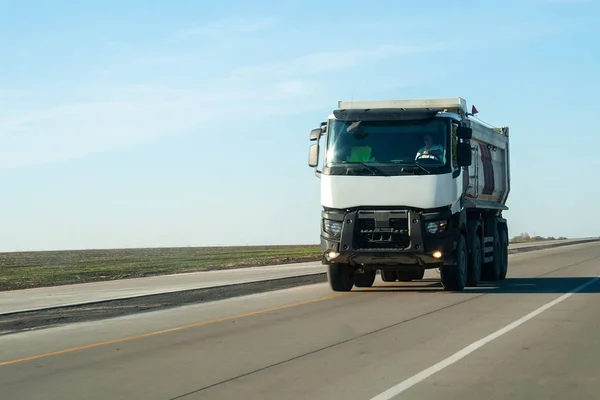 Biała wywrotka wyjeżdża z miasta na płaskiej asfaltowej drodze wśród pól na tle błękitnego nieba w słoneczny dzień. Maszyna do transportu materiałów budowlanych na autostradzie — Zdjęcie stockowe