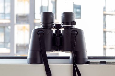 Pencerenin bulanık arka planına karşı siyah dürbünler komşu evi, çevreyi ya da doğayı gözlemlemek için görüş açısına sahip. Seçici odaklanma. Yakın görünüm