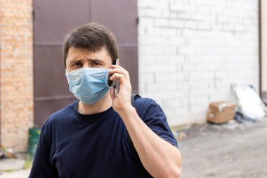 Beyaz bir adamın sokak portresi. Salgın Coronavirus covid-19 döneminde, koruyucu mavi bir tıbbi maskeyle akıllı telefondan konuşan bir adam. Virüse karşı korunmak için tek kullanımlık solunum cihazındaki erkek.