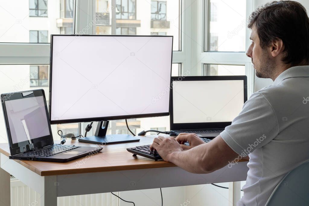 A man in a white t-shirt is sitting at a workplace with two laptops and a monitor near the window. Remote work from home office