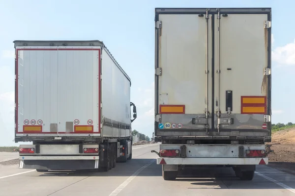 Güneşli bir günde bulutlu mavi gökyüzüne karşı düz bir asfalt yolda ilerleyen iki damperli kamyonun arka görüntüsü. Şehirlerarası bir otoyolda yol çalışır.