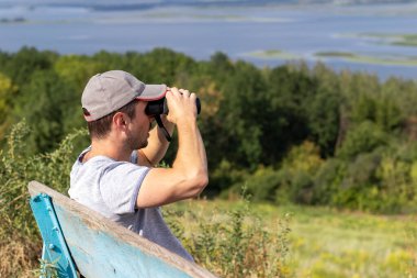 Bir adam, tepedeki eski ahşap bir bankta otururken geniş bir nehrin manzaralı manzarasına dürbünle bakar. Yan görünüm