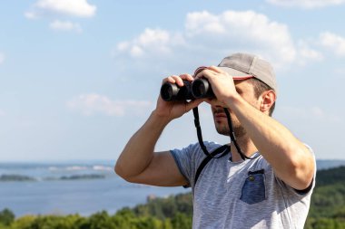 Dürbünle bakan bir adam güneşli bir günde geniş bir nehrin manzarasına bakar.