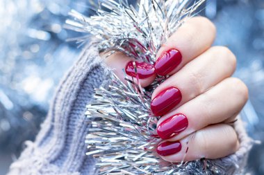 Gri örgü süveterli, güzel, parlak manikürlü bir kadın eli. Bordo, koyu kırmızı, kiraz rengi tırnaklar. Gümüş Noel süslerinin arka planında. Seçici odaklanma. Yakın görünüm