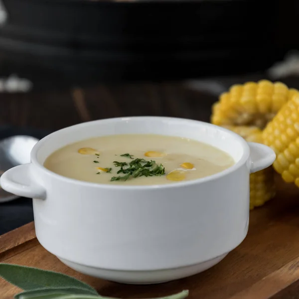 Close view of corn soup in bowl with fresh corn cob on black plate.