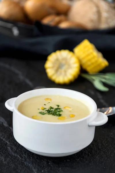 Close view of corn soup in bowl with fresh corn cob on black plate.