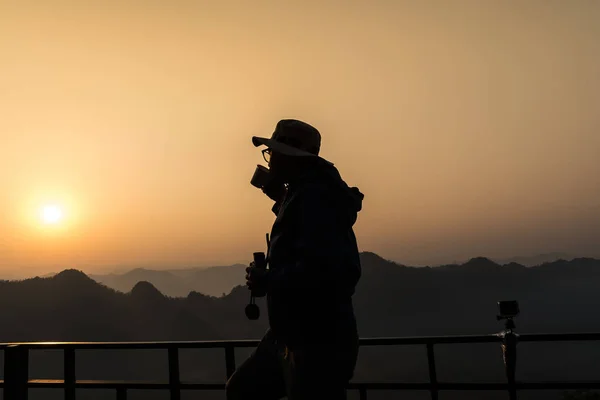 Silhouette of photographer drinking tea in morning, mountains on background.