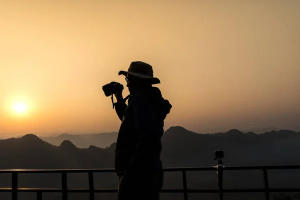 Silhouette of photographer in hat looking at mountains with binoculars in morning.