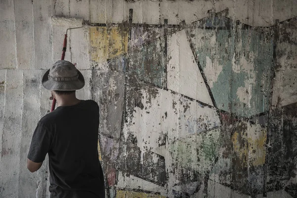 Young man in hat cleaning walls by painting white over old cement wall, Thailand.