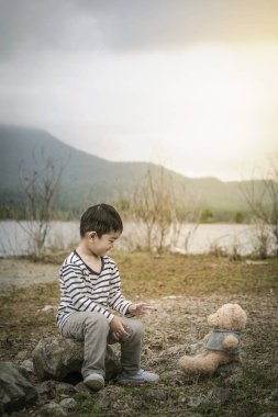 Oyuncak ayı ile rezervuar arka plan üzerinde taşlar üzerinde oturan Asya çocukla.