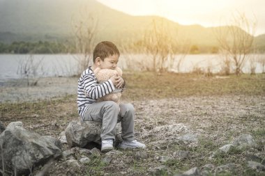 Asyalı çocuk rezervuar üzerinde arka plan ile taşlar üzerinde oturan bir oyuncak ayı ile sarılma.