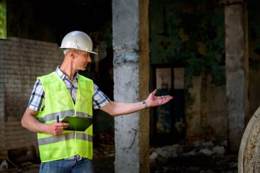 İnşaat kaskı olan ve elinde siyah bir klasör olan bir adam inşaat alanında. Bir çalışma şeklinde tuğgeneral, yapının incelenmesi.