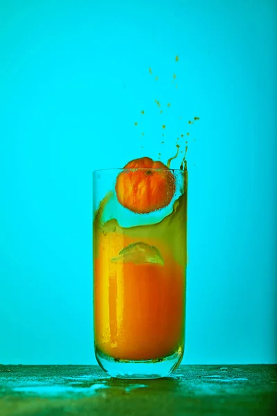 Une tranche d'orange tombant dans un verre avec du jus d'orange et un soupçon de jus sur un fond bleu. Concept de saine alimentation — Photo