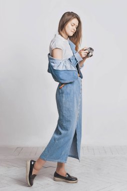 Fotoğraf stüdyosunda beyaz arka planda güzel bir kız. Retro kamera tutuyor. Kot pantolon giyiyordu. Denim uzun etek ve ceket. Nostalji 90 'lar.