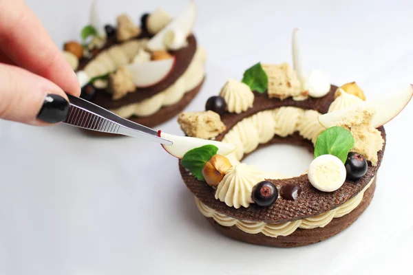 Hand with tweezers finishing rustic Paris Brest choux pastry rings with hazelnut ganache, nuts, apple slices, black currants and hazelnut sponge — Stock Photo, Image