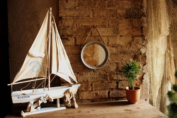 Small decorative yacht on table with potted plant and round mirror hanging on a red brick wall