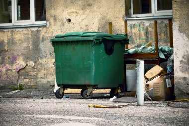 Kapalı kapaklı yeşil plastik çöp konteynırı pencereli gri bina duvarının önünde duruyor.