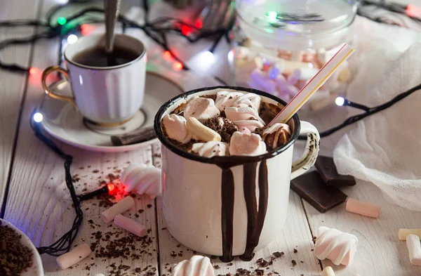 Mug of hot chocolate drink with marshmallow candies on top and candles and Christmas lights.