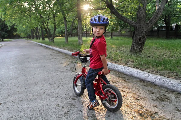 A small white Toddler boy in a protective helmet on his head sits on a children\'s bicycle. Toddler on a two-wheeled red bicycle looks back. A sly smile on the child\'s face
