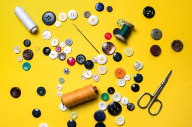 Dikiş takımı. Çok renkli plastik düğmeler, iplik makaraları, dikiş iğneleri ve makas parlak sarı bir arka planda yatar.
