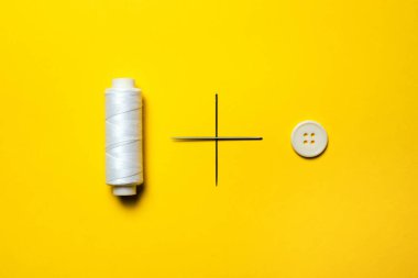 Beyaz iplik makarası, artı işareti oluşturan nakış iğneleri ve parlak sarı arka planda beyaz bir düğme var.
