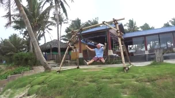 Vietnam Hanoi december 20, 2018: lycklig ung man i skjorta och shorts Swinging på en sväng på en ö i Vietnam — Stockvideo