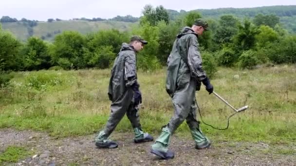 Dos cadetes militares de un soldado en el campo de Chernóbil están buscando minas con un detector de minas bajo la lluvia. Protegido en un traje de peligro de radiación — Vídeo de stock