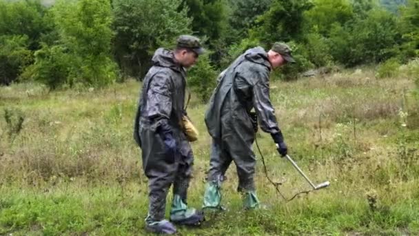 チェルノブイリの森林地帯の兵士の2人の軍幹部は、雨の中で鉱山探知機を持つ鉱山を探しています。放射線の危険からスーツで保護される — ストック動画