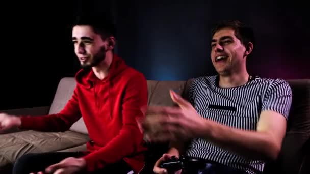 Émotionnel deux jeunes gars sont assis sur le canapé en face de la télévision et excité de jouer à une console de jeu, tenant un joystick. Jeux pour hommes avec une manette sans fil. Chambre confortable est éclairée avec lumière au néon . — Video