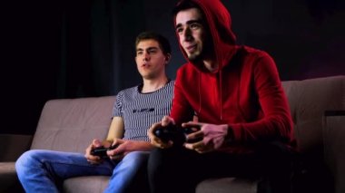 Duygusal iki genç adam kanepede oturup oyun konsolu oynayıp, elinde joystick tutarak heyecanlılar. Kablosuz kontrolörle oyun oynayan erkekler. Sade oda neon ışıklarıyla aydınlatılmış..