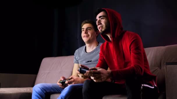 Emocional dois jovens estão sentados no sofá na frente da TV e animado jogando um console de jogo, segurando um joystick. Mens jogos com um controlador sem fio. Quarto acolhedor é iluminado com luz de néon . — Vídeo de Stock