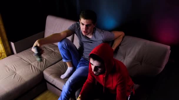 İki genç adam TV 'nin karşısında kanepede oturmuş oyun konsolu oynuyor, elinde joystick tutuyorlardı. Bir kablosuz kontrolörle oynarken insanlarla iletişim kurmak. Sade oda neon ışıklarıyla aydınlatılmış.. — Stok video