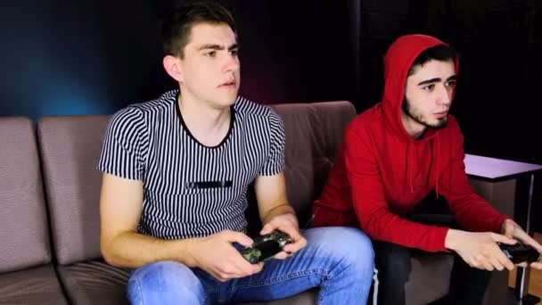 Dois jovens estão sentados no sofá na frente da TV e jogando um console de jogo, segurando um joystick. Comunicando mans enquanto joga com um controlador sem fio. Quarto acolhedor é iluminado com luz de néon . — Vídeo de Stock