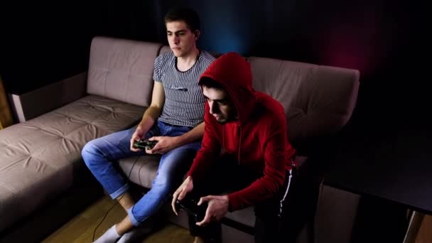 Twee jonge jongens zitten op de bank voor de TV en spelen een spelcomputer met een joystick. Communiceren met mensen tijdens het gamen met een draadloze controller. Gezellige kamer is verlicht met neon licht. Stockvideo's