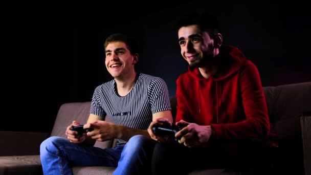 Dwóch młodych facetów się śmieje. Chłopcy siedzący na kanapie przed telewizorem i grający na konsoli do gier, trzymający joystick.Communicating mans podczas gry z kontrolerem bezprzewodowym.Pokój oświetlony jest neonowym światłem — Wideo stockowe