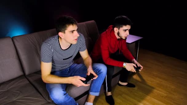 Émotionnel deux jeunes gars sont assis sur le canapé en face de la télévision et excité de jouer à une console de jeu, tenant un joystick. Jeux pour hommes avec une manette sans fil. Chambre confortable est éclairée avec lumière au néon . — Video