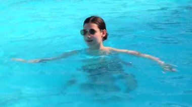 Yüzme havuzunda güneş gözlüklü güzel esmer kız. Mavi havuz suyunda mayo giymiş bir kadın lüks otellerde yaz yolculuğunun tadını çıkarıyor. Güneşli yaz gününde parlak güneş ışığı yansımalarıyla.