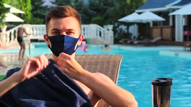 Homme caucasien agitant la main regarder la caméra. Jeune homme bronzant avec masque médical de protection montrent bonjour signal de la main. Le concept de monde du coronavirus met en quarantaine l'épidémie Covid-19. Fond de piscine. — Video