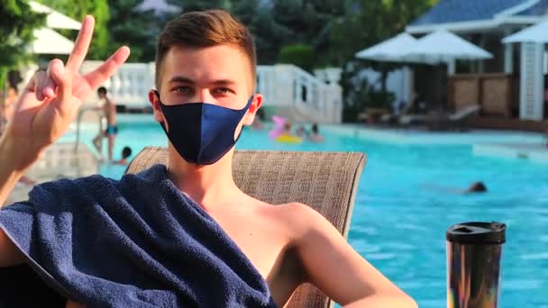 Homme caucasien montre le geste de paix regarder la caméra. Jeune homme bronzant avec masque médical de protection montrent bonjour signal de la main. Épidémie de Coronavirus Covid-19. Fond de piscine. — Video