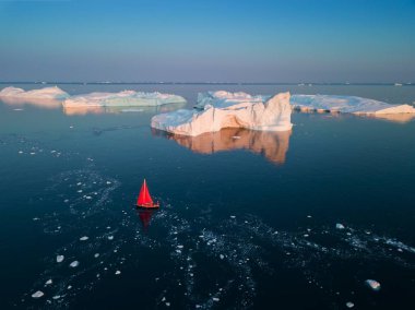 Little red sailboat cruising among floating icebergs in Disko Bay glacier during midnight sun season of polar summer. Ilulissat, Greenland. Arctic nature ice landscape in Unesco World Heritage Site.  clipart